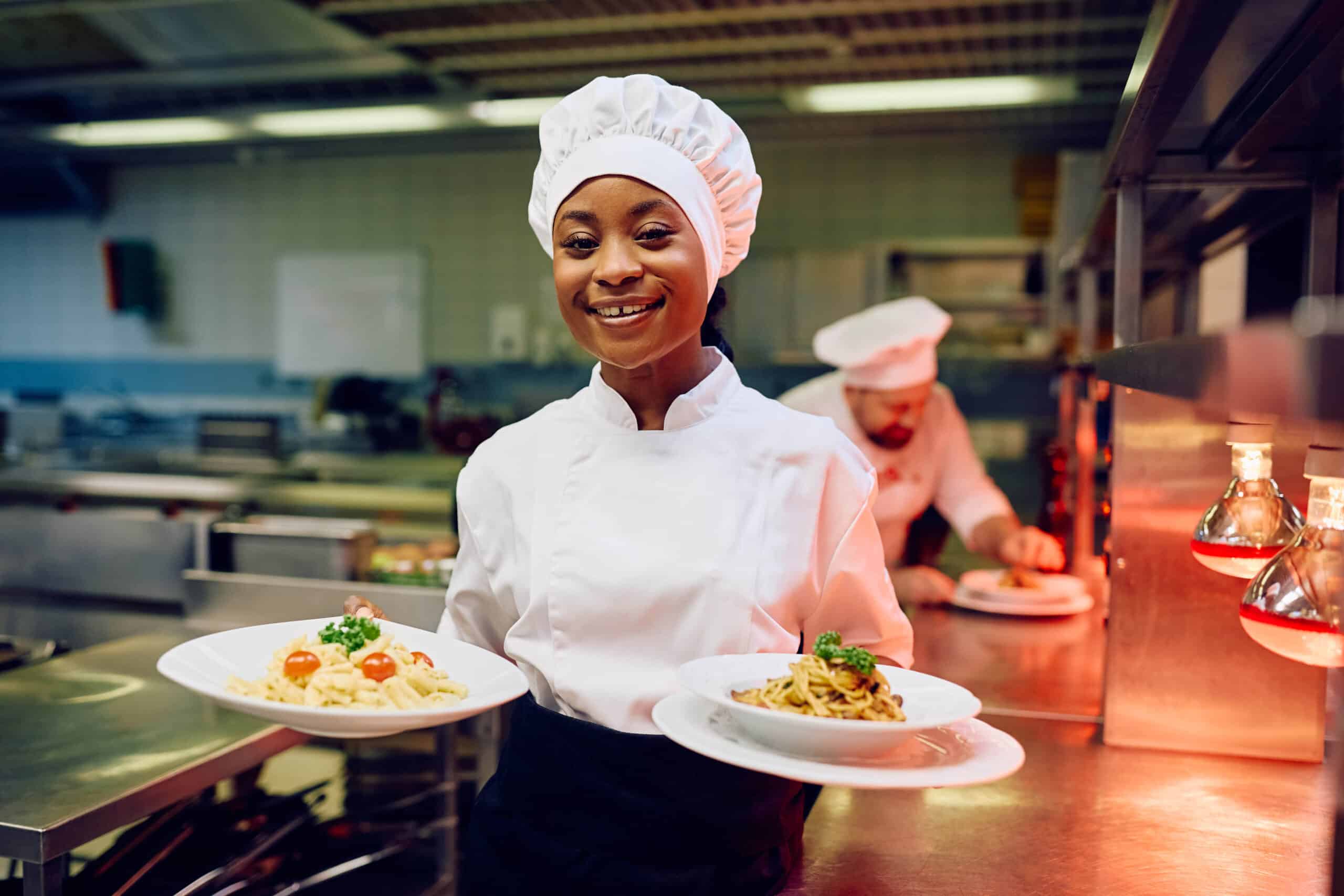 Mala Barad smiling and holding two pasta dishes in a professional kitchen